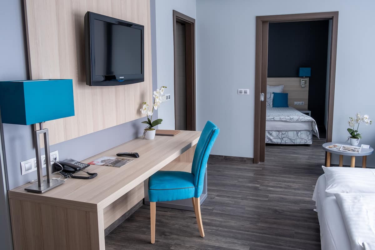 A hotel room with a wooden desk, a blue chair, a bed, a TV, and a small table with a lamp.