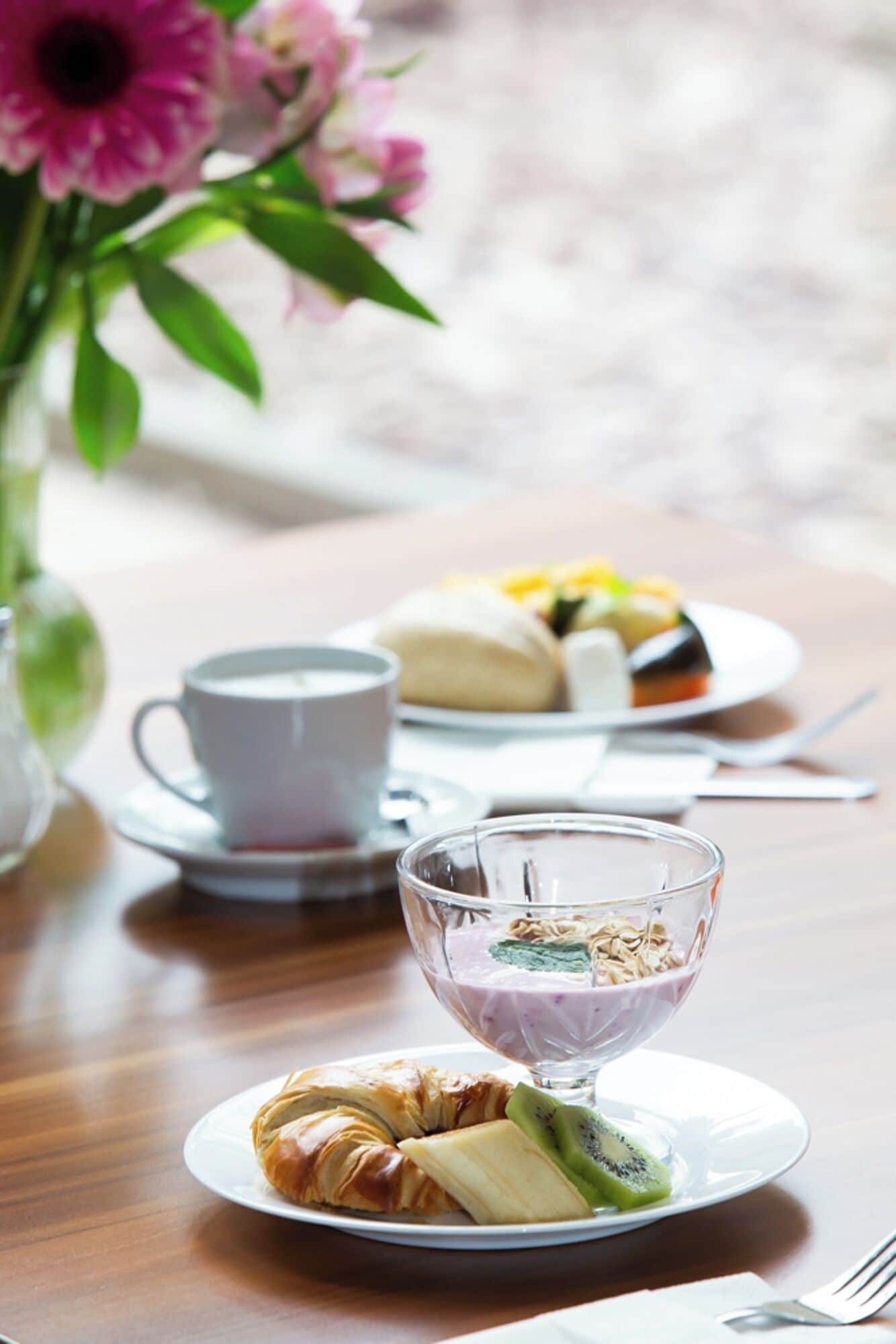 A table with a croissant, a glass bowl of yogurt with fruit, a cup of coffee, and a plate of food.