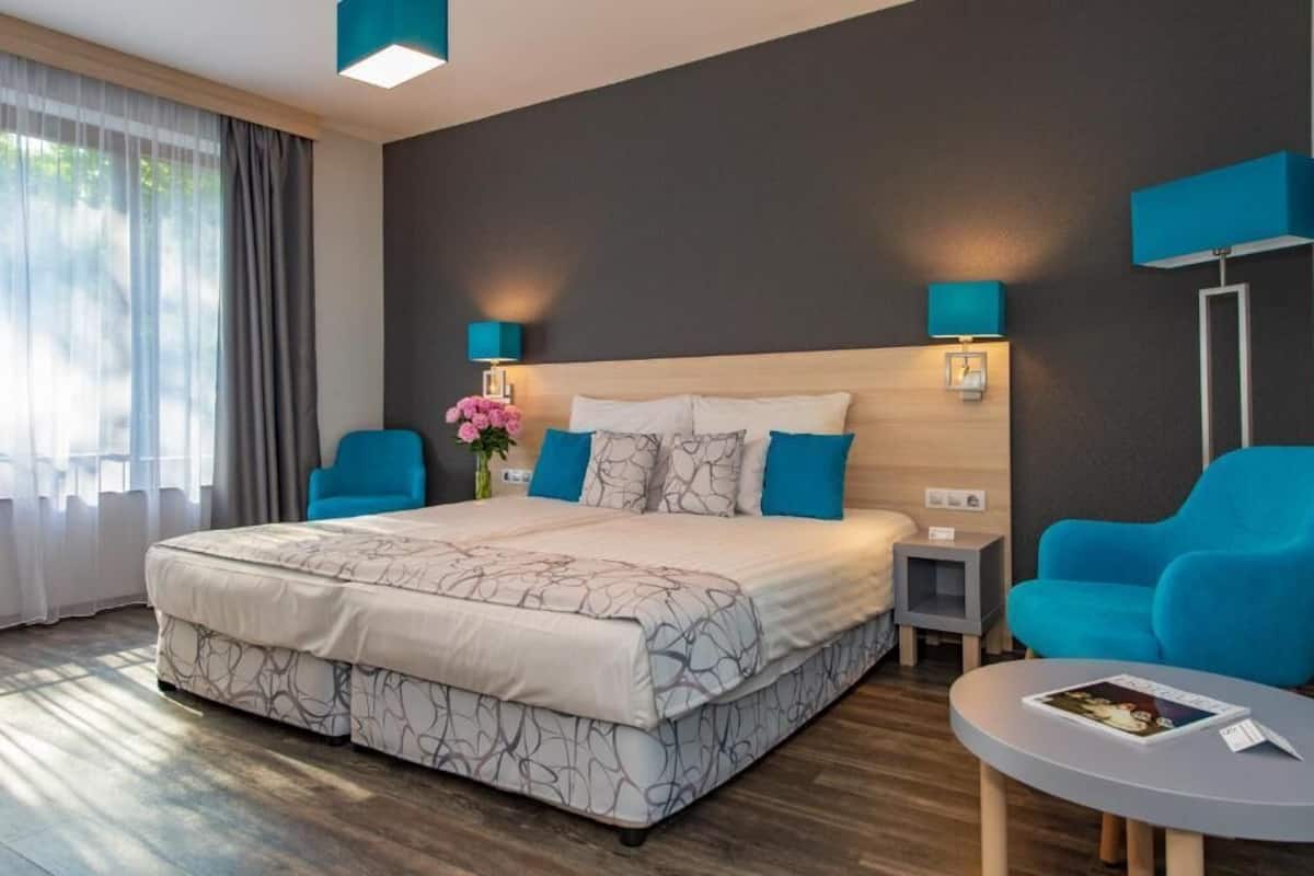 A modern hotel room with a large bed, two blue armchairs, a small table, and a nightstand.