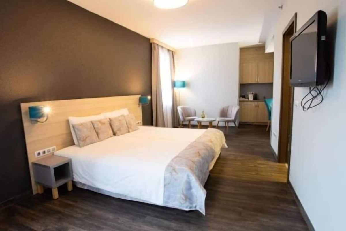 A hotel room with a bed, bedside table, a TV mounted on the wall, and a small kitchenette area.