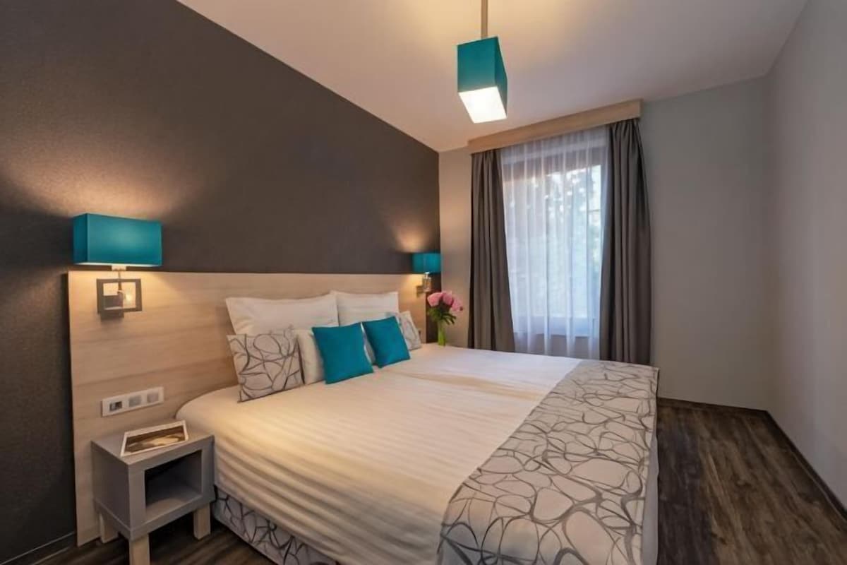 Hotellihuone, jossa on suuri sänky, yöpöydällä olevat lamput, yöpöytä ja ikkuna, jossa on verhot.
