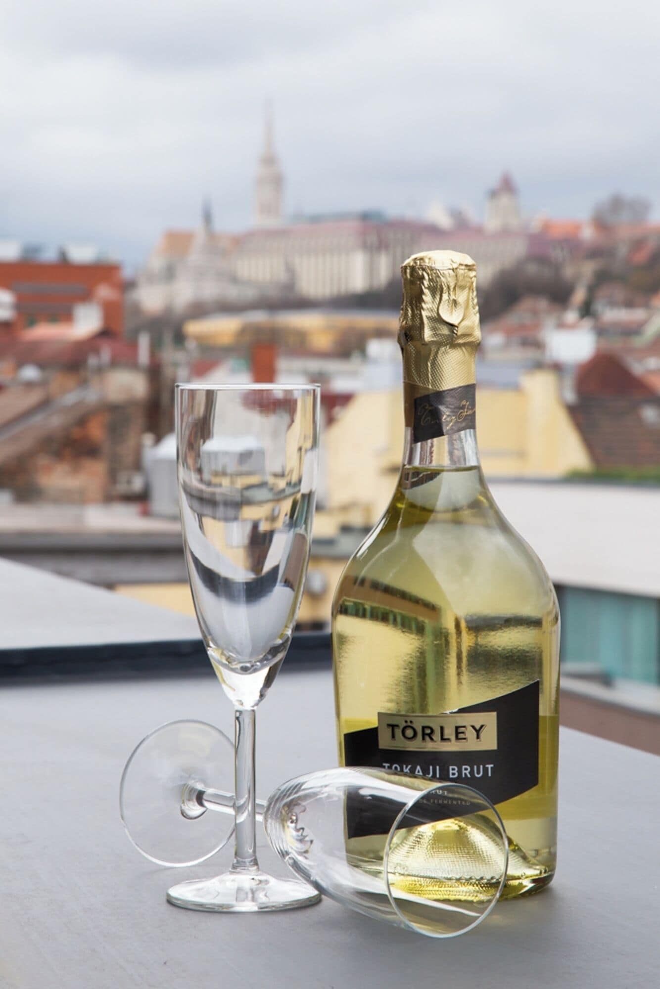 A bottle of Tokaji Brut wine and a wine glass on a table with a cityscape view in the background.