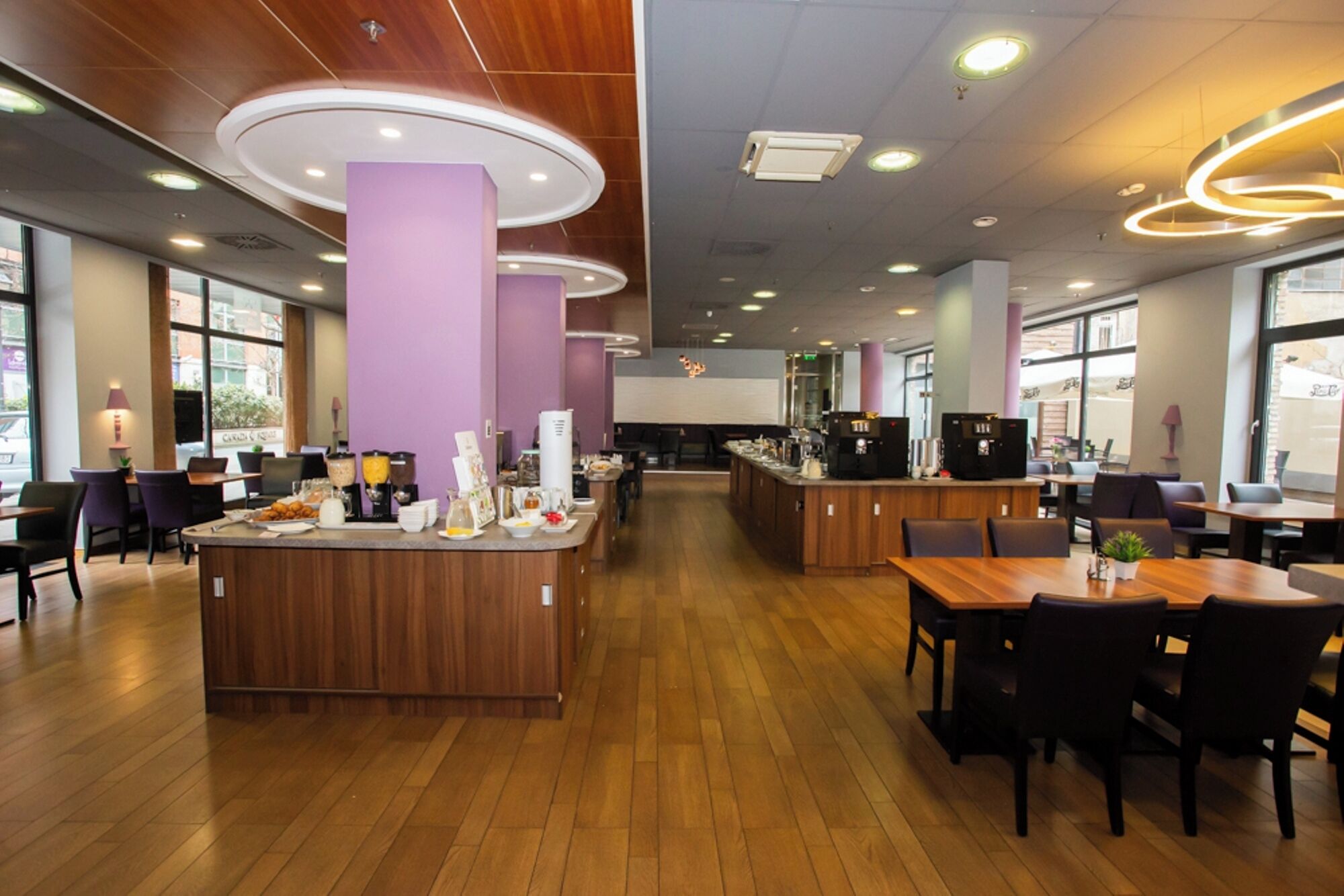 Ein modernes Café mit Holzböden, violetten Akzenten und einer zentralen Theke mit verschiedenen Speisen und einer Kaffeemaschine.