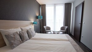 Une chambre d’hôtel avec un grand lit, deux chaises, une petite table, une table de chevet et une fenêtre avec des rideaux.