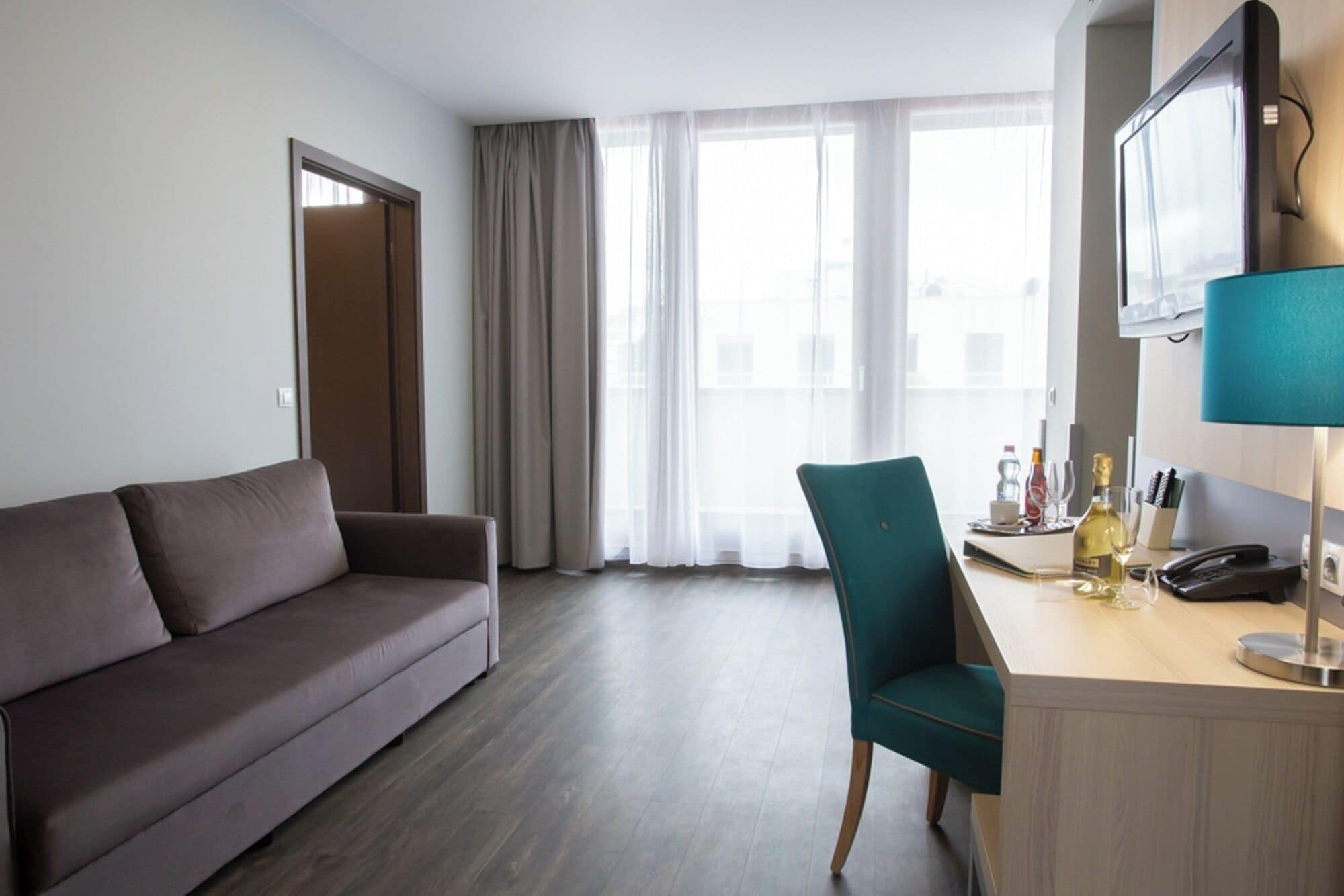 Ein Hotelzimmer mit grauem Sofa, einem türkisfarbenen Sessel, einem Holztisch mit Telefon, einer Weinflasche und einem Fernseher auf einem Standfuß.