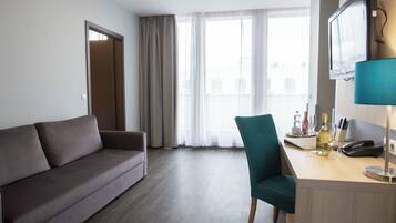 Ein Hotelzimmer mit grauem Sofa, einem türkisfarbenen Sessel, einem Holztisch mit Telefon, einer Weinflasche und einem Fernseher auf einem Standfuß.