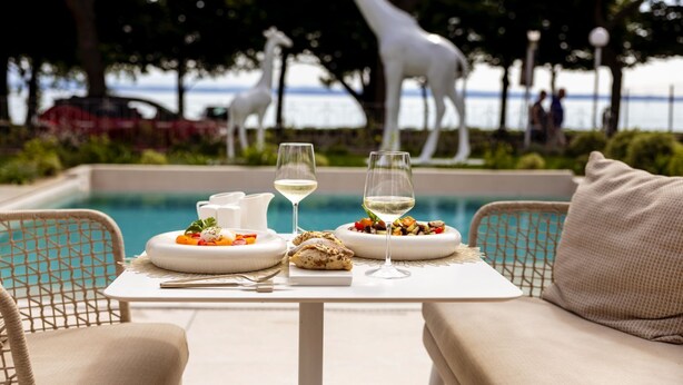 Una mesa de comedor con platos de comida y copas de vino, una estatua de jirafa blanca y una piscina al fondo.