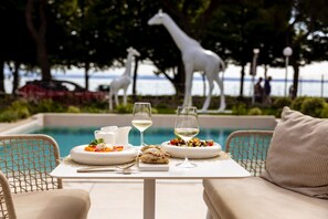 Una mesa de comedor con platos de comida y copas de vino, una estatua de jirafa blanca y una piscina al fondo.