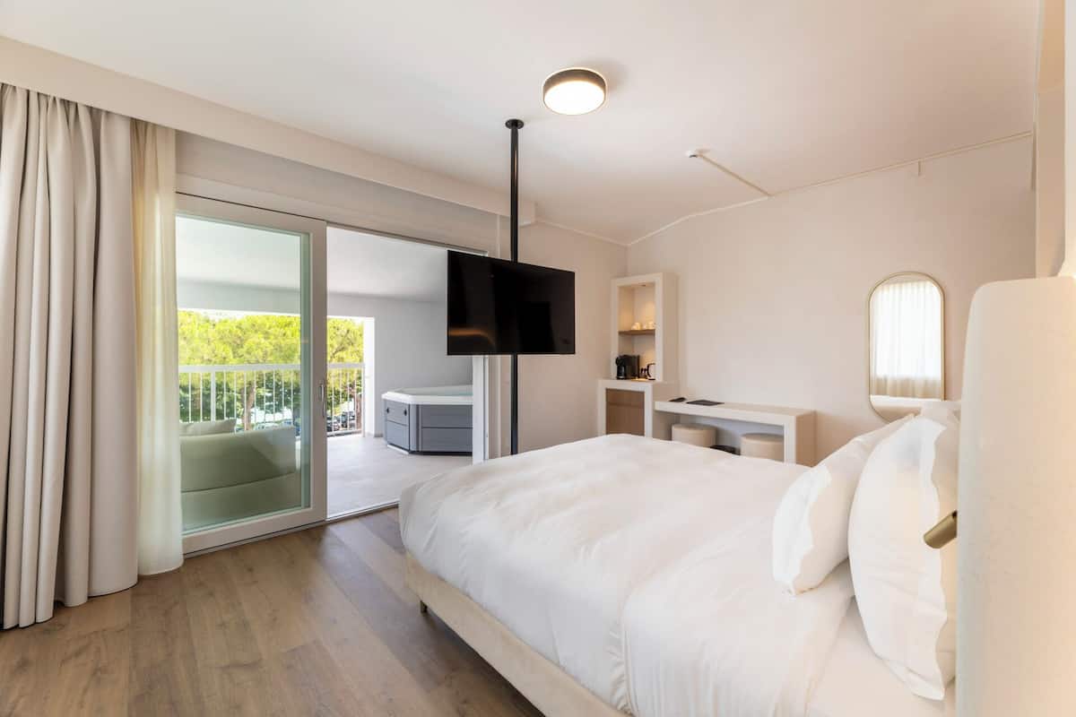 A modern hotel room with a large bed, a flat-screen TV, a desk, and a balcony with a view of greenery.