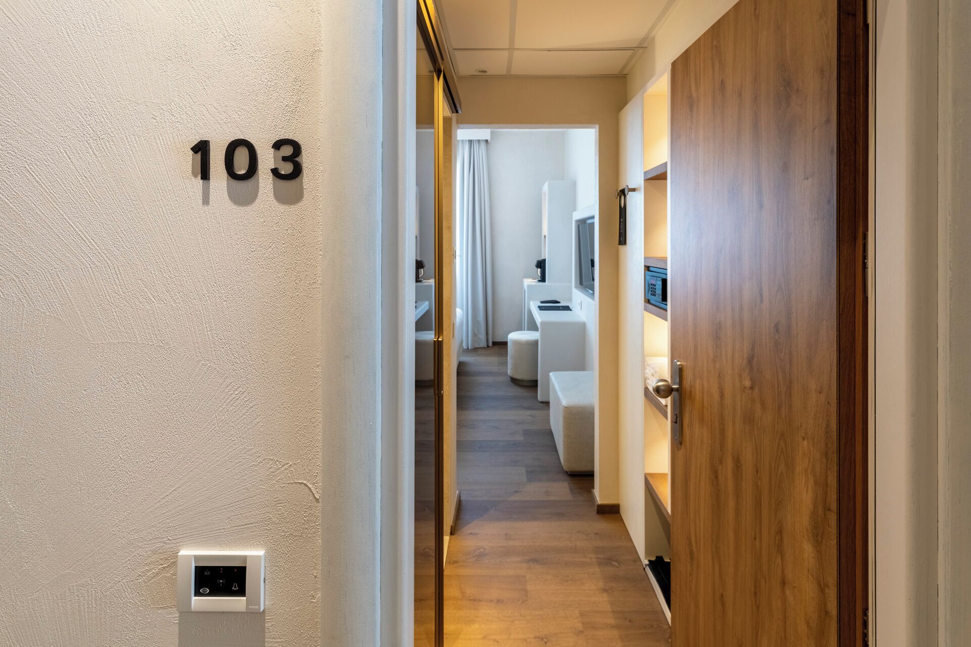 Een gang met het nummer '103' op de muur, die leidt naar een kamer met een houten deur en een witte muur.