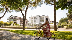 Una mujer pedalea en bicicleta por un camino pavimentado, con árboles y un edificio al fondo.