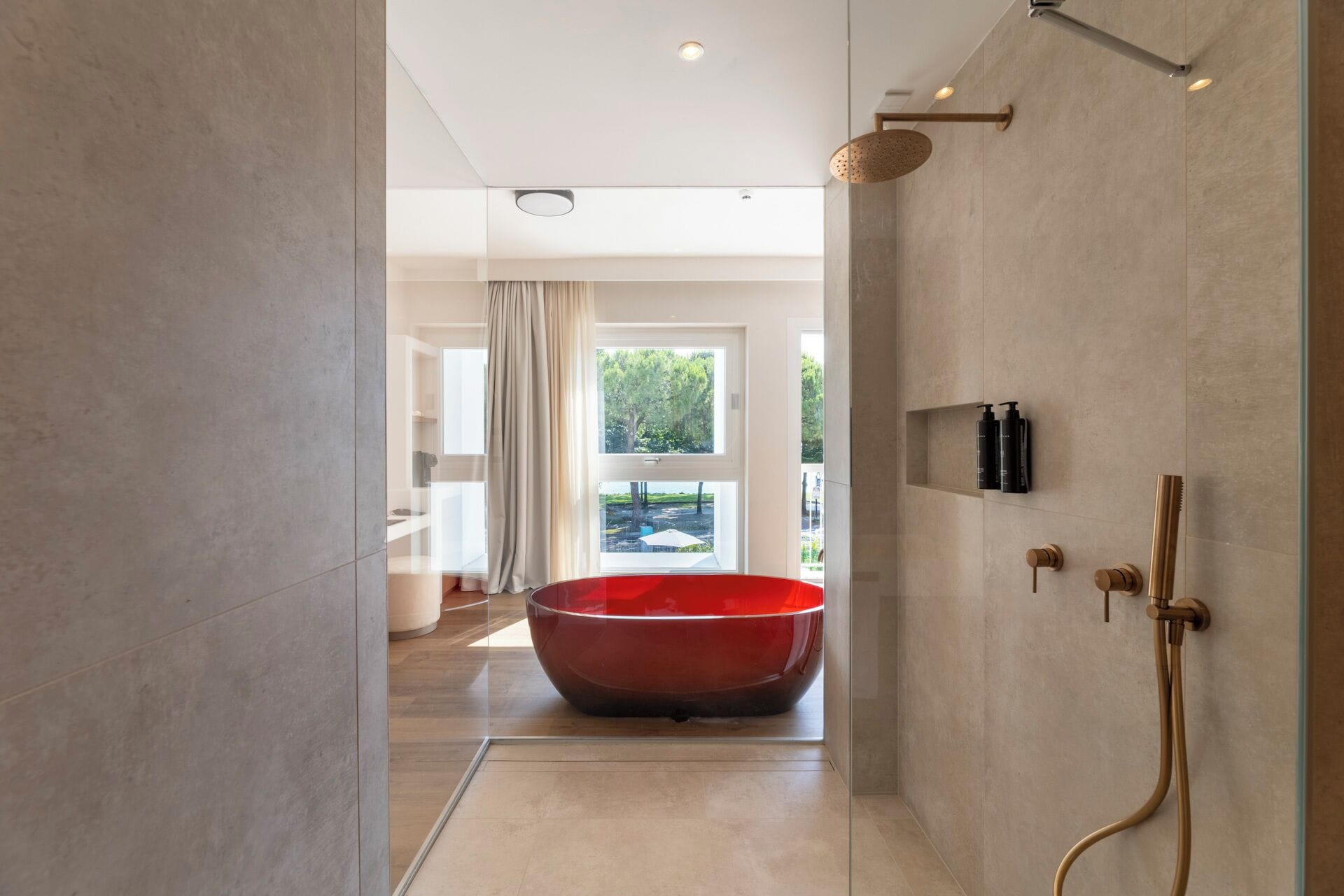 A modern bathroom with a large red bathtub, a shower area with gold fixtures, and a view of a garden through a large window.