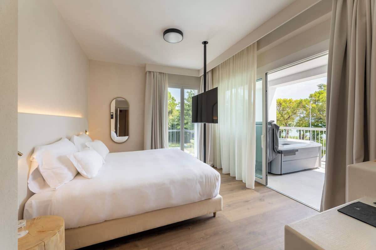 A hotel room with a large bed, a flat-screen TV, a balcony with a view of trees, and a wooden floor.