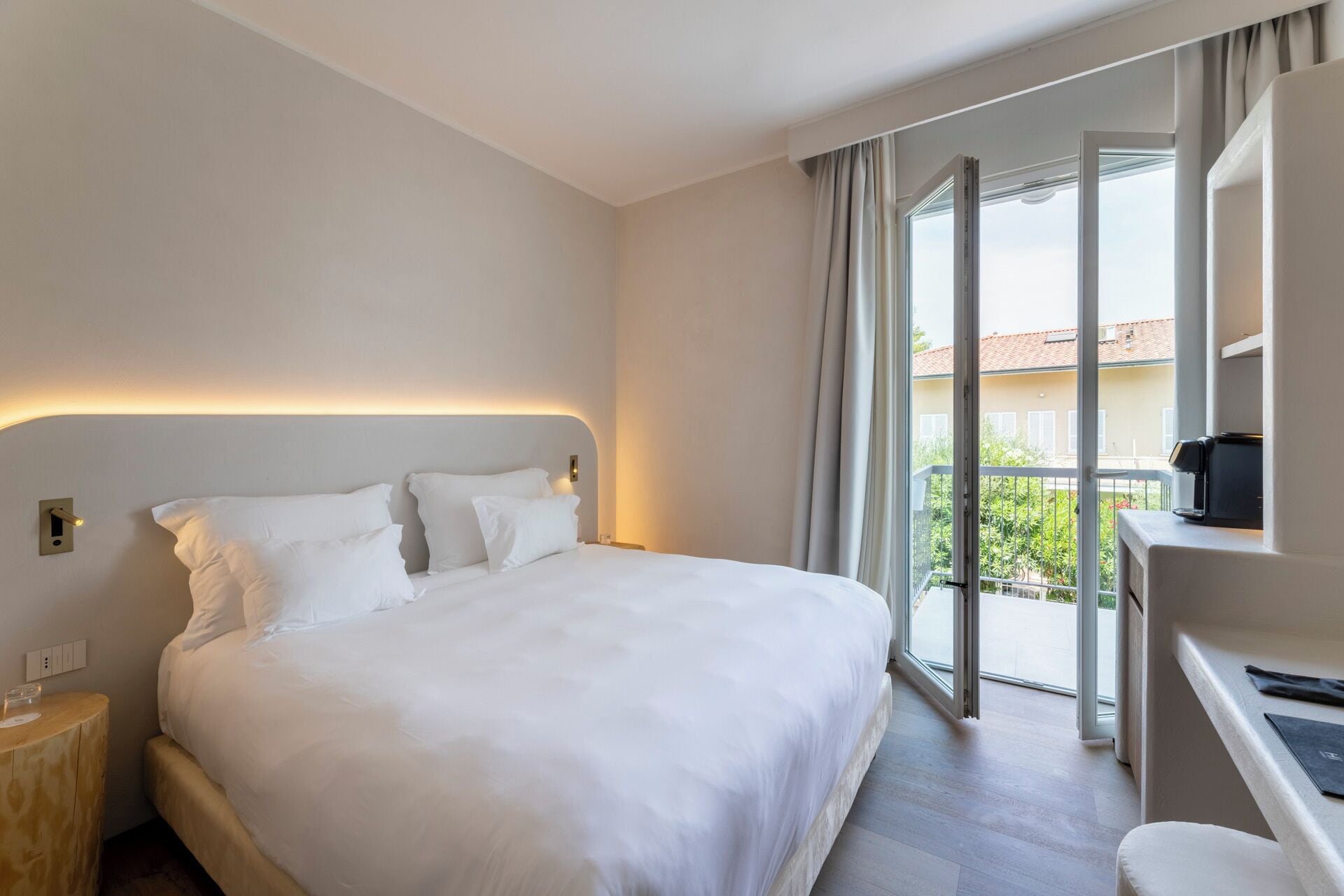 A hotel room with a large bed, a bedside table, a desk with a chair, and a balcony with a view of the outside.