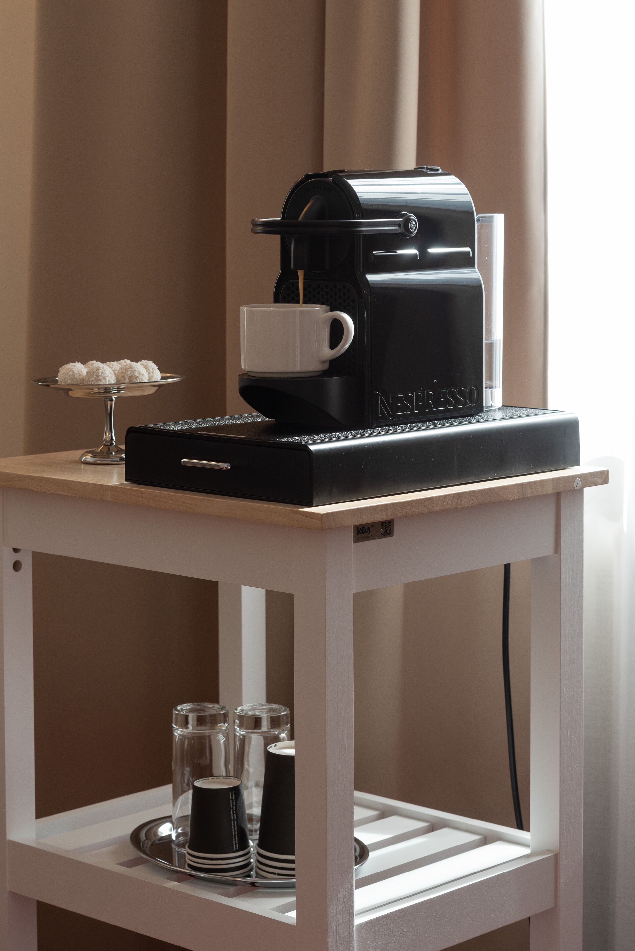 Una máquina de café Nespresso sobre una mesa blanca, con una bandeja que contiene tazas y una jarra de vidrio.