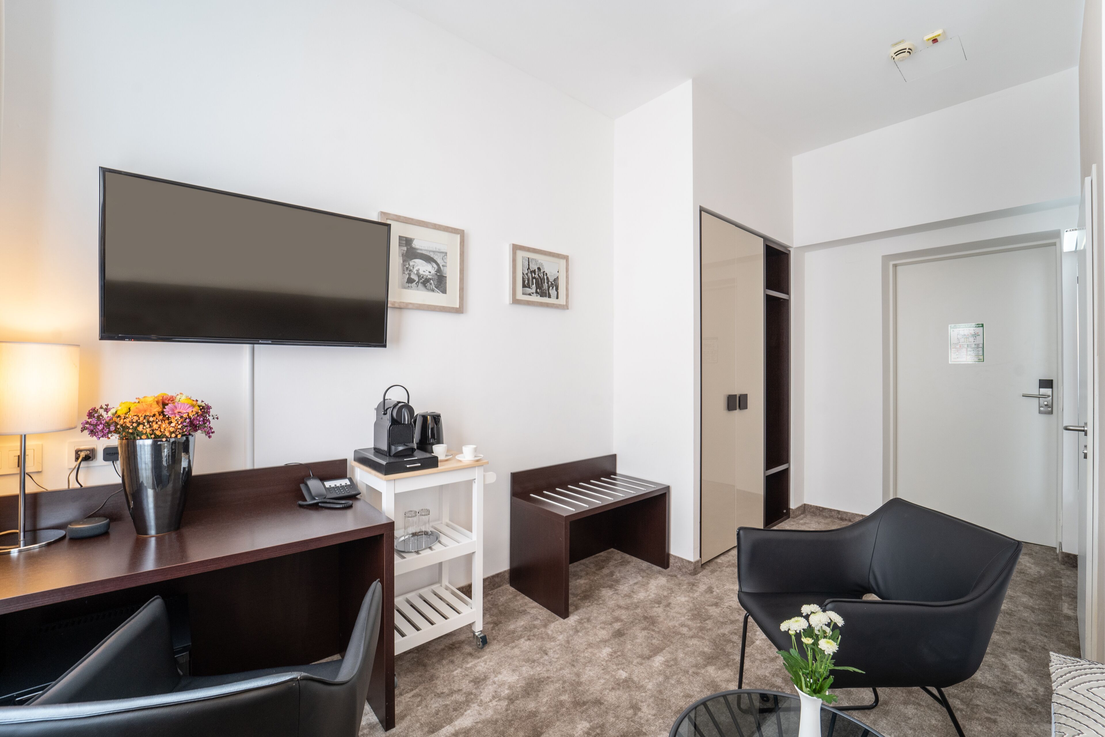 Et hotelværelse med skrivebord, stol, TV og et lille bord med blomster.