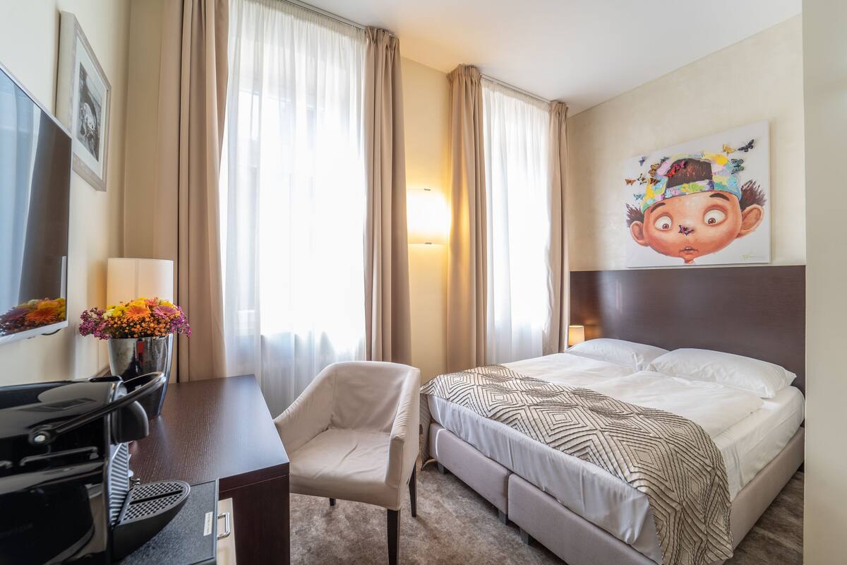 Una camera d'albergo con un letto, una scrivania con una lampada, una sedia e un quadro appeso al muro.