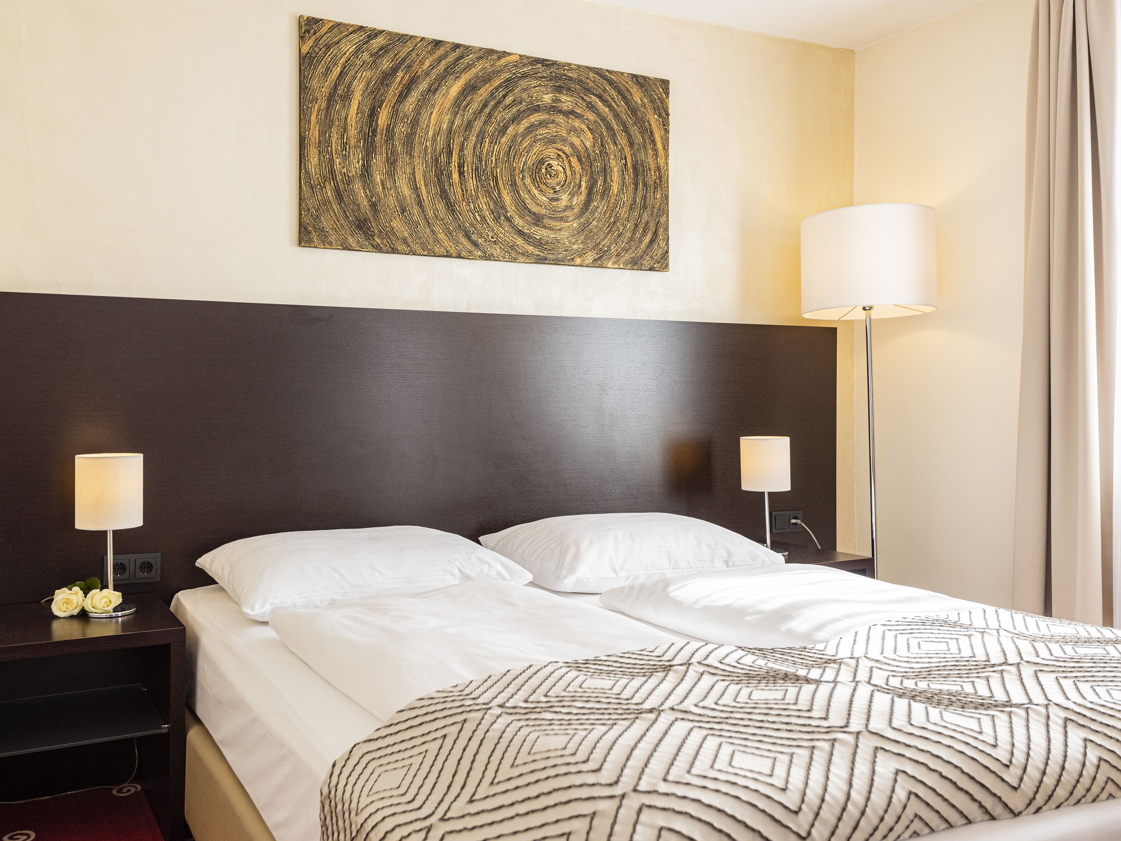 A hotel room with a large bed, two bedside tables, a wall-mounted artwork, and two floor lamps.