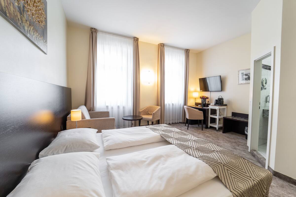 Una camera d'albergo con un letto grande, una scrivania, una sedia, una TV e un bagno visibile attraverso una porta aperta.