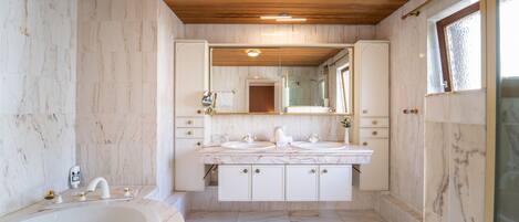 A bathroom with marble walls and floor, a wooden ceiling, a large bathtub, and a double sink vanity with cabinets.