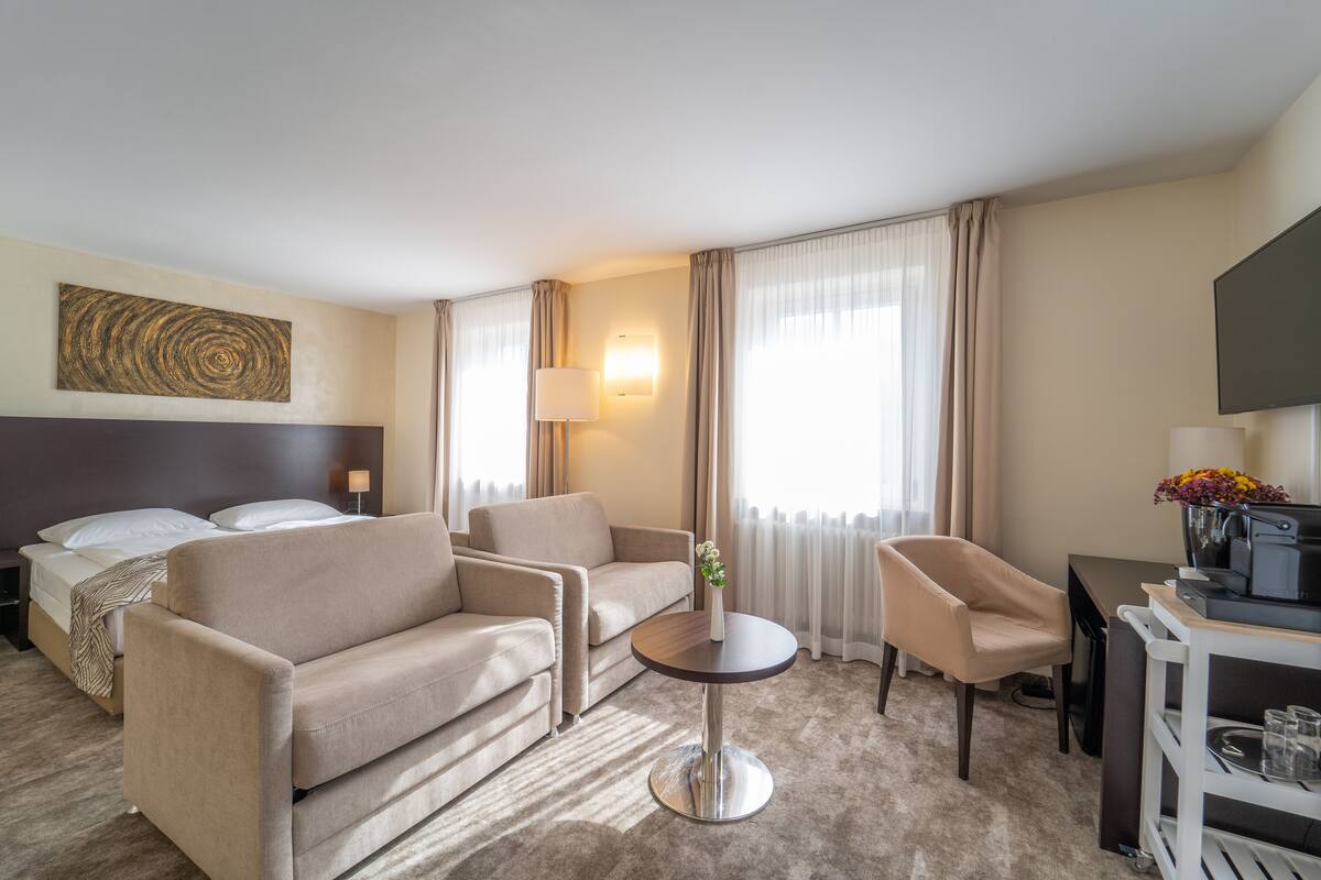Una camera d'albergo con un letto, due divani, un tavolino rotondo, una scrivania con un computer, una TV e un quadro grande appeso al muro.