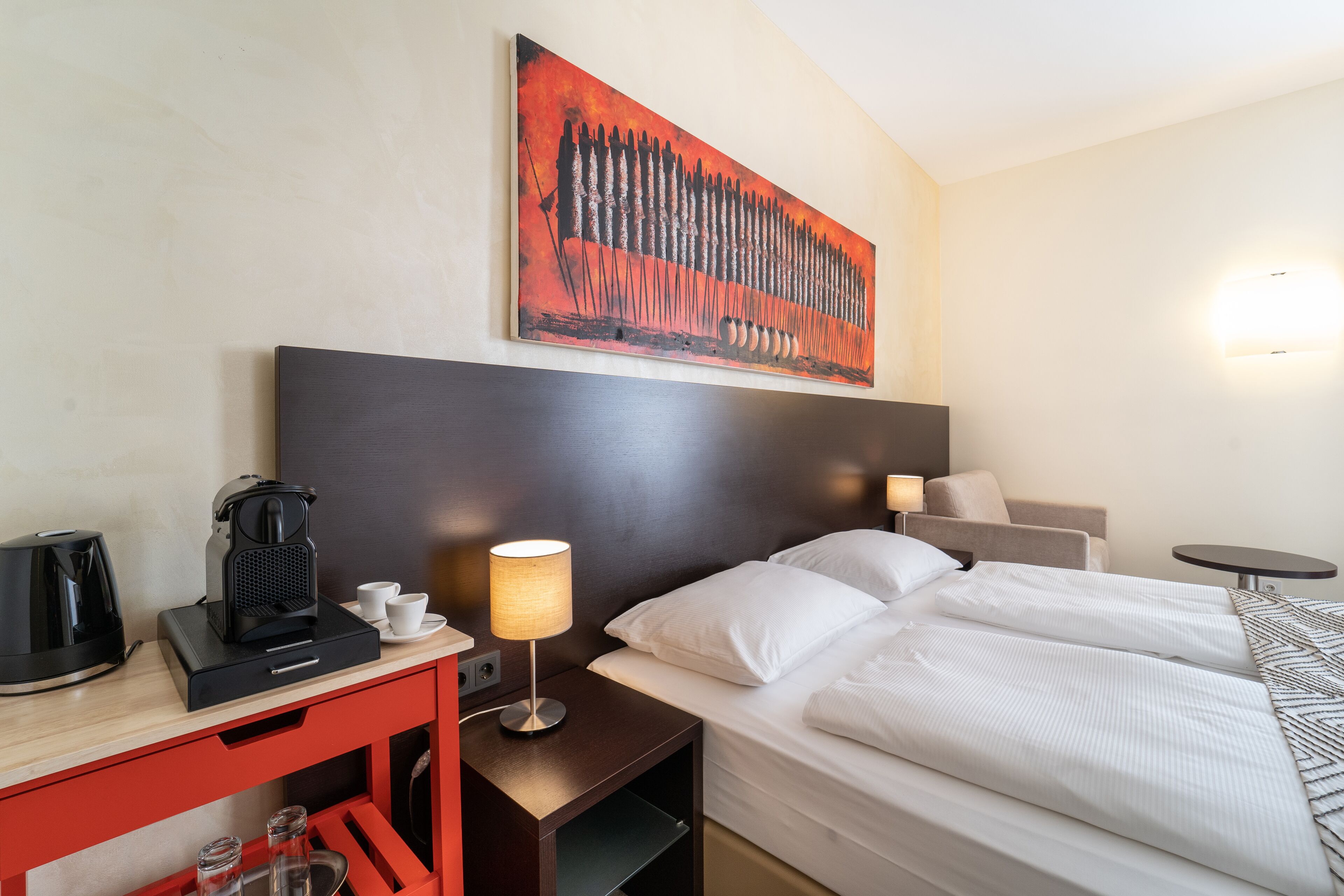 A hotel room with a bed, bedside table with a lamp, a coffee maker, and a red cabinet.