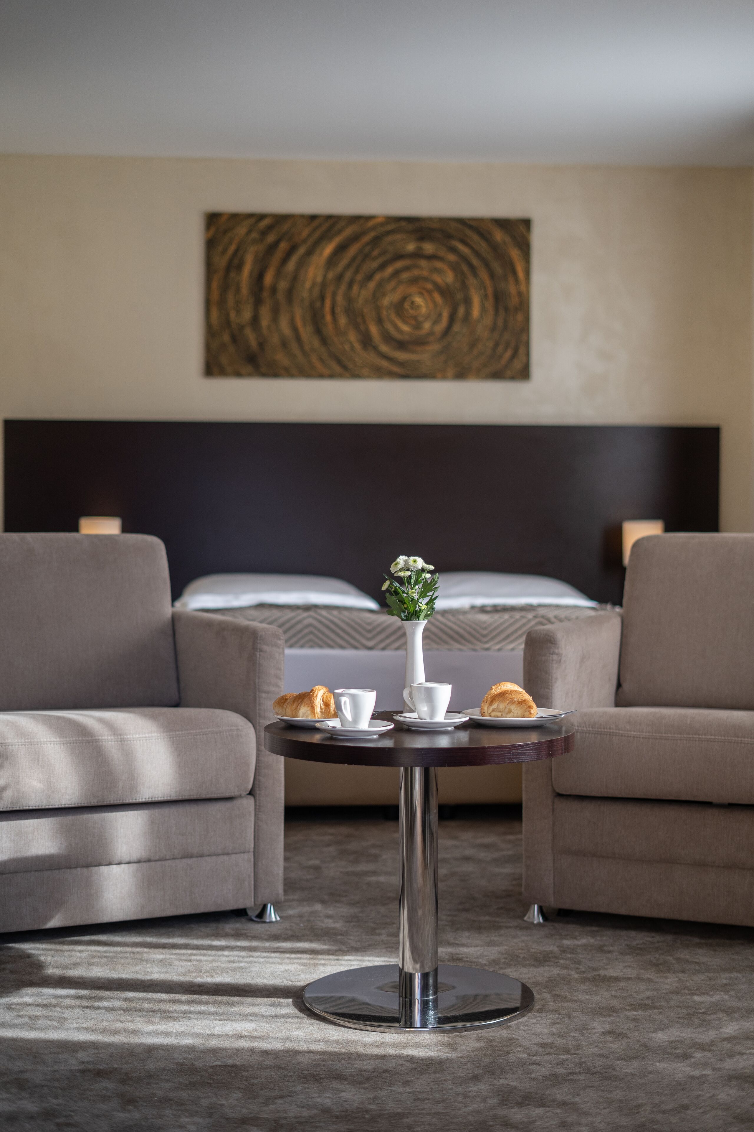 A hotel room with a bed, two armchairs, a small round table with a vase of flowers, and a coffee cup.