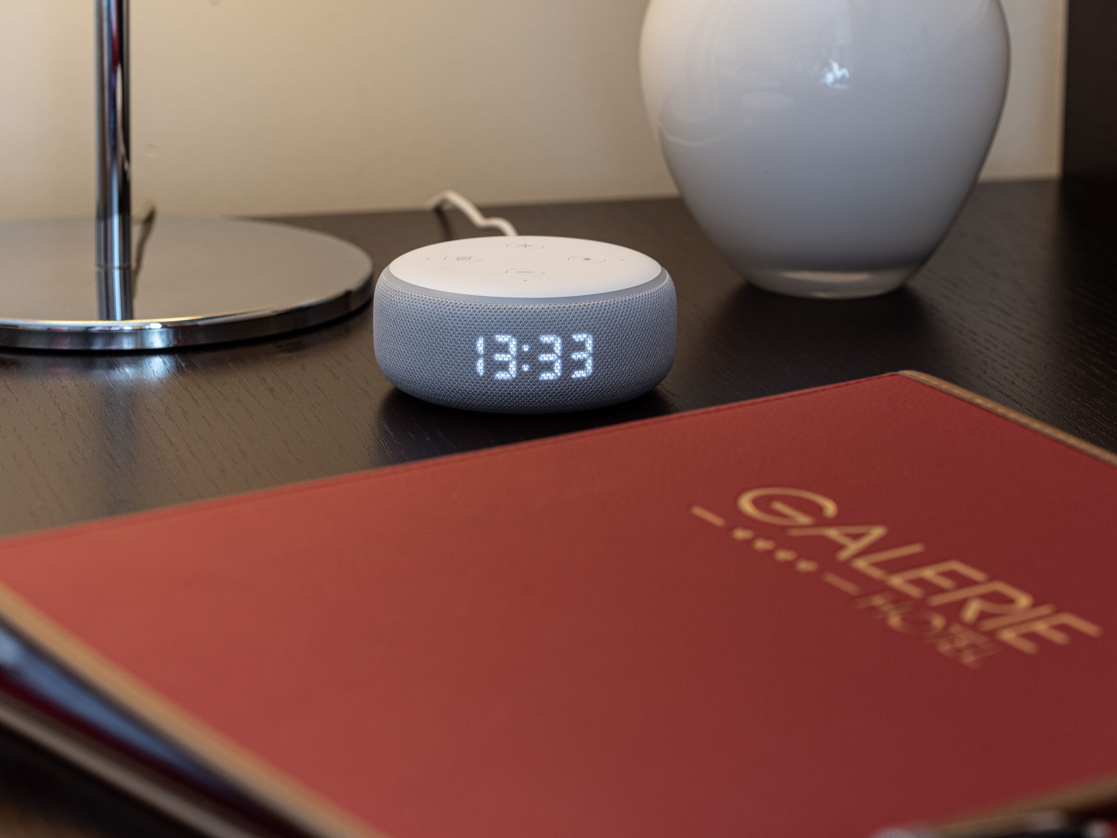 A smart speaker on a table with a red book titled "GALERIE" and a white vase.