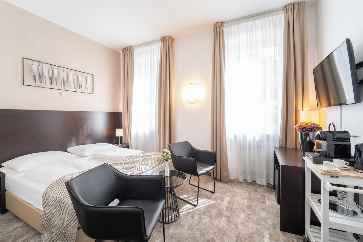 Una camera d'albergo con un letto, due sedie nere, un tavolino, una televisione, una scrivania con una macchina da caffè e una lampada a parete.