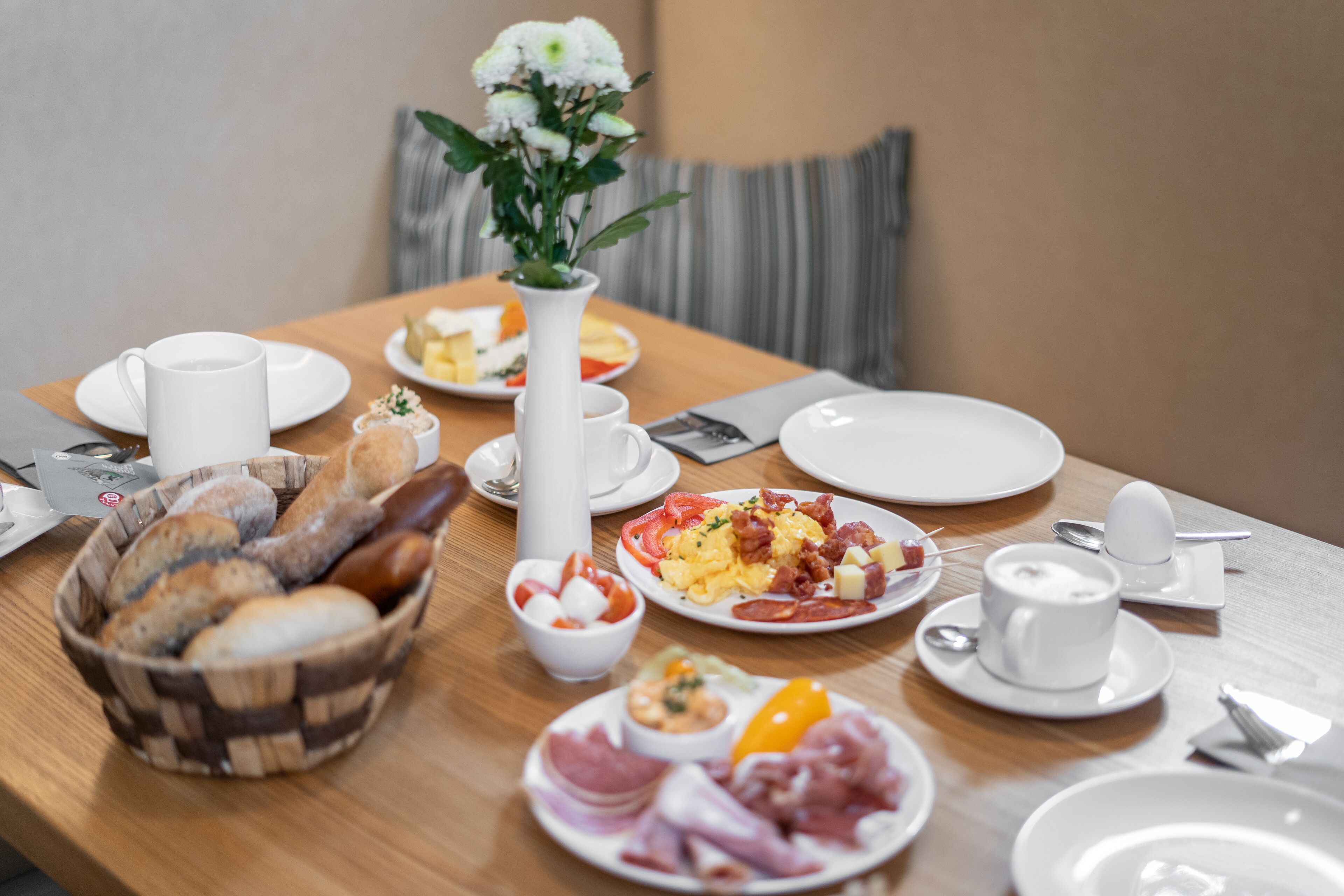 A breakfast table with various dishes, a white vase with flowers, and a striped cushion in the background.