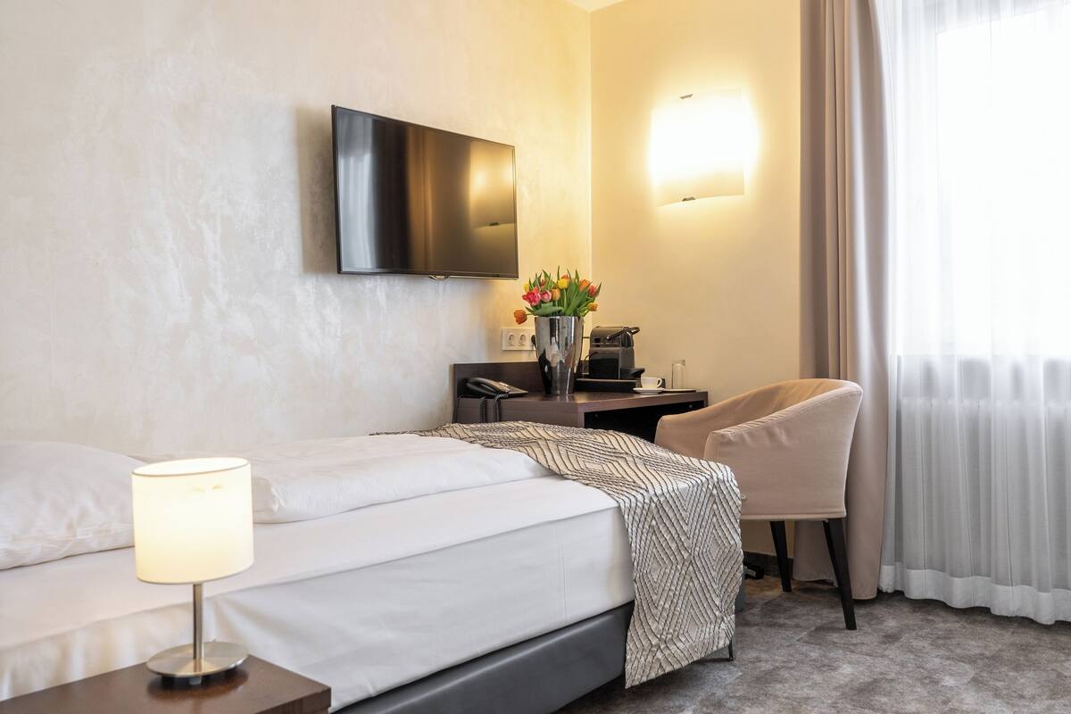 Una camera d'albergo con un letto, una scrivania con una lampada, una sedia, una TV a parete e una finestra con tende.