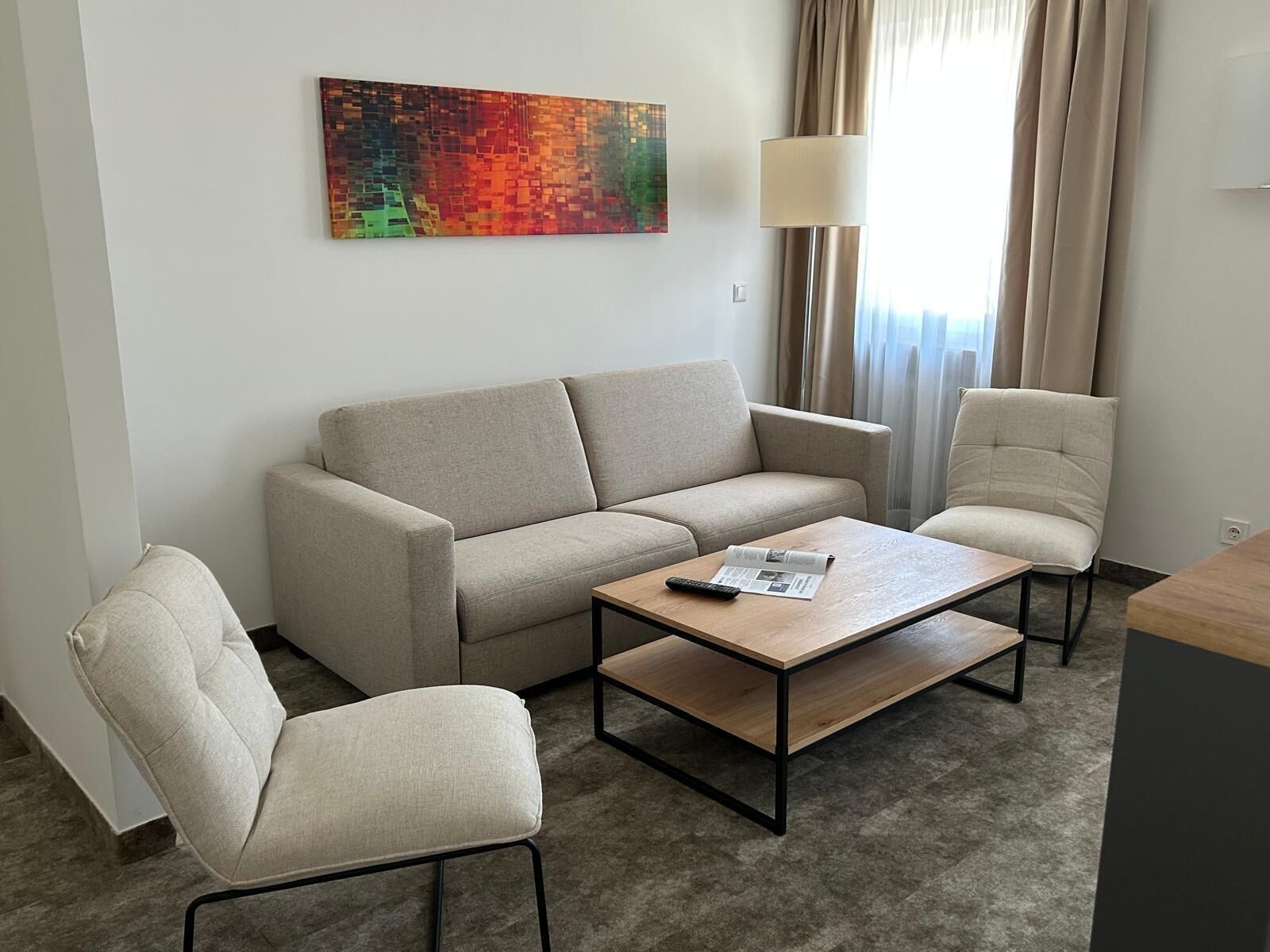 Une chambre d’hôtel moderne, dotée d’un canapé, d’un fauteuil, d’une table basse et d’un tableau abstrait aux couleurs vives accroché au mur.