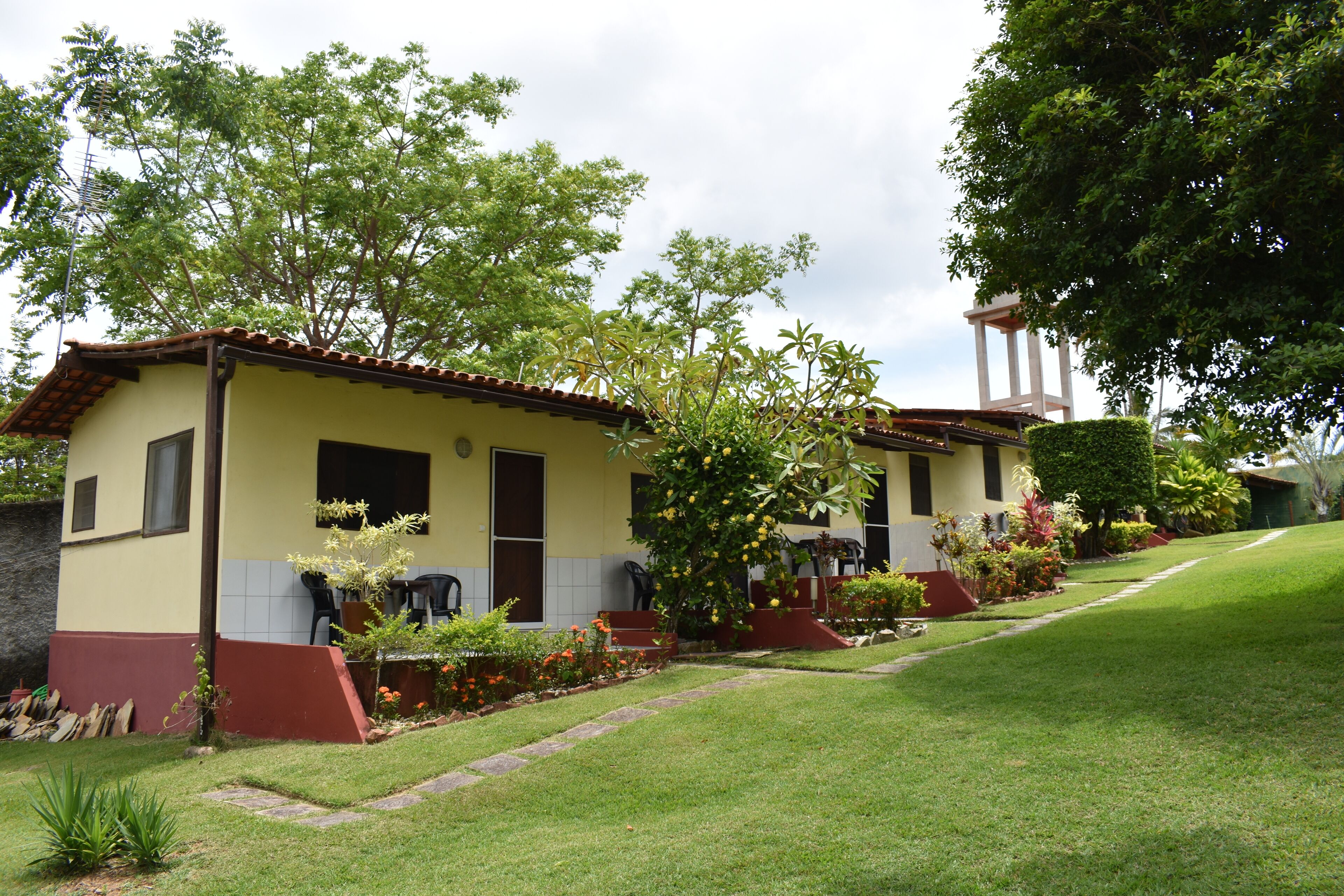 Uma fileira de edifícios de dois andares com fachada em vermelho e amarelo, cercada por jardins bem cuidados e diversas plantas.