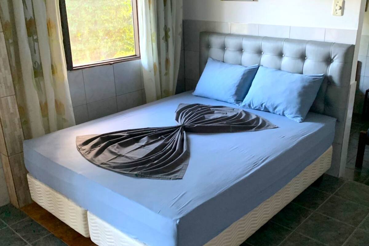Un lit bien fait, avec une couvre-lit bleue et deux oreillers, une fenêtre avec des rideaux et une tête de lit blanche.