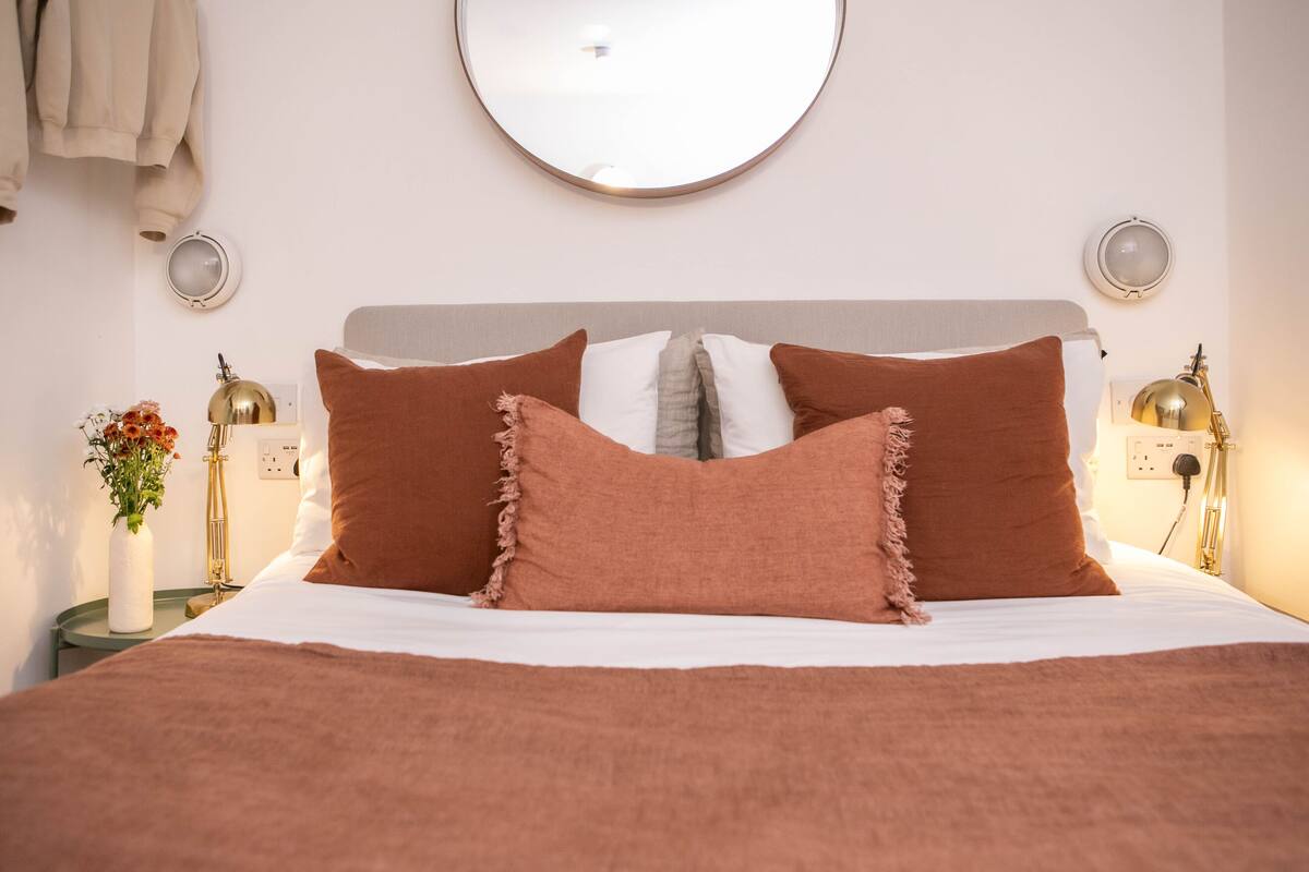 Una cama bien hecha con almohadas marrones y un edredón blanco, un espejo redondo en la pared y lámparas de noche.