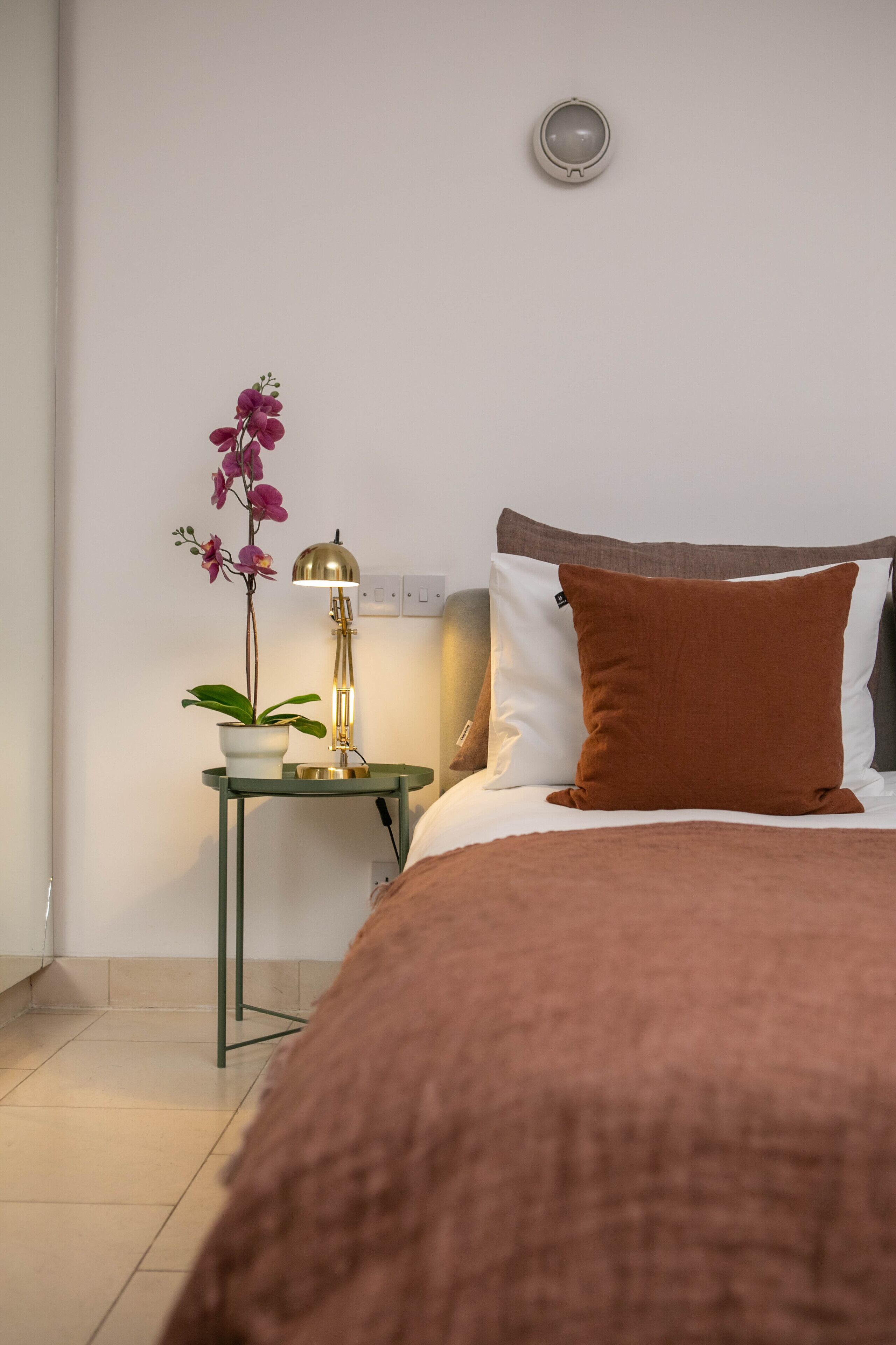 Un dormitorio ordenado con una cama, una mesita de noche con lámpara y una orquídea en maceta.