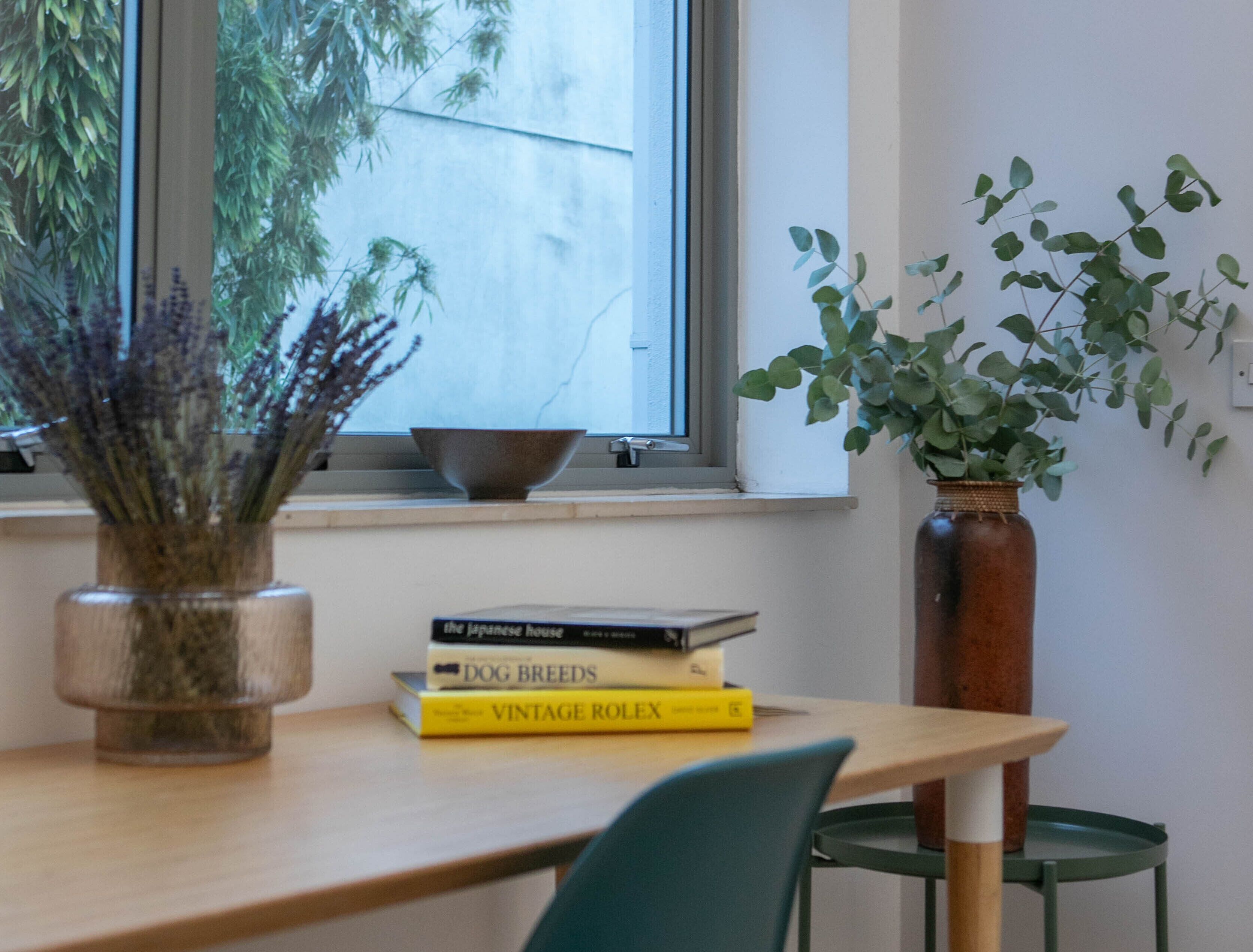 A room with a window, a bowl on the sill, a vase with plants, and a stack of books on a table.