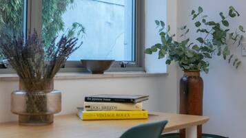 A room with a window, a bowl on the sill, a vase with plants, and a stack of books on a table.