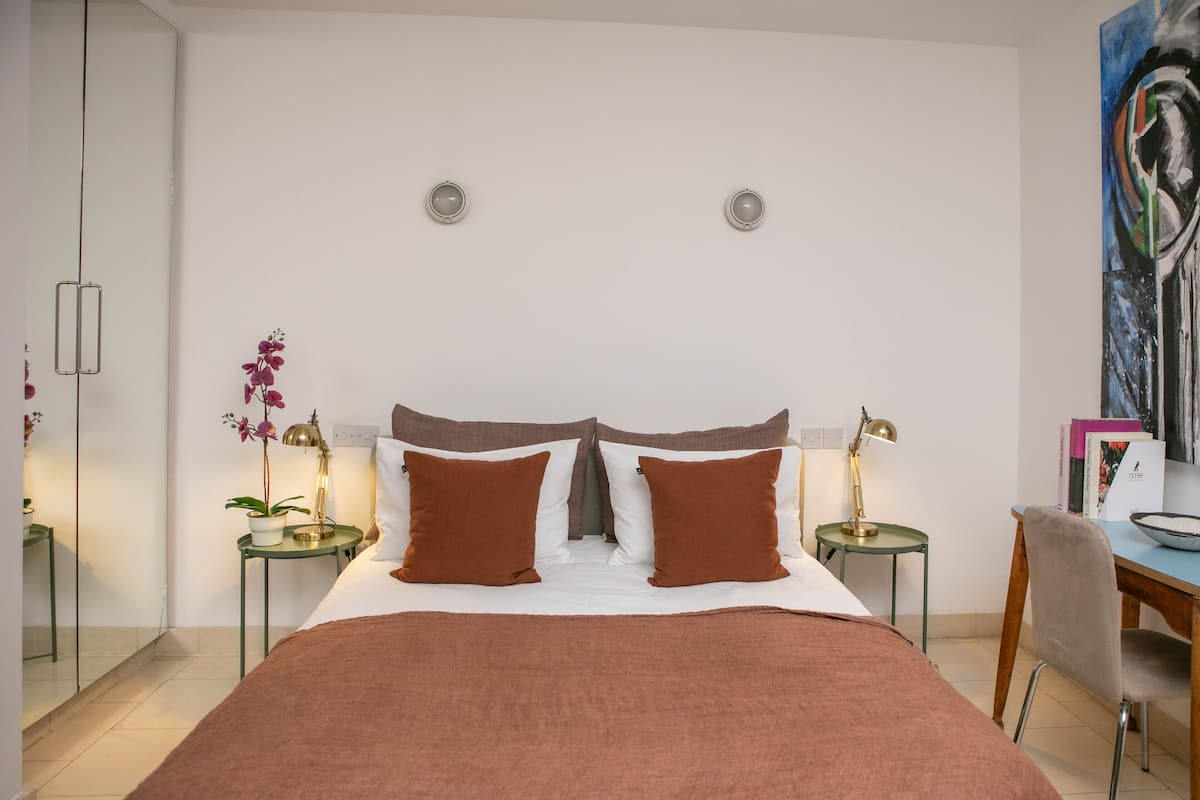 Una habitación de hotel moderna con una cama grande, dos mesitas de noche con lámparas, un escritorio con una silla y un cuadro abstracto de gran tamaño en la pared.