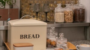 Una encimera de cocina con un recipiente para pan, botes de cereales, un plato de cruasanes y una taza de café.