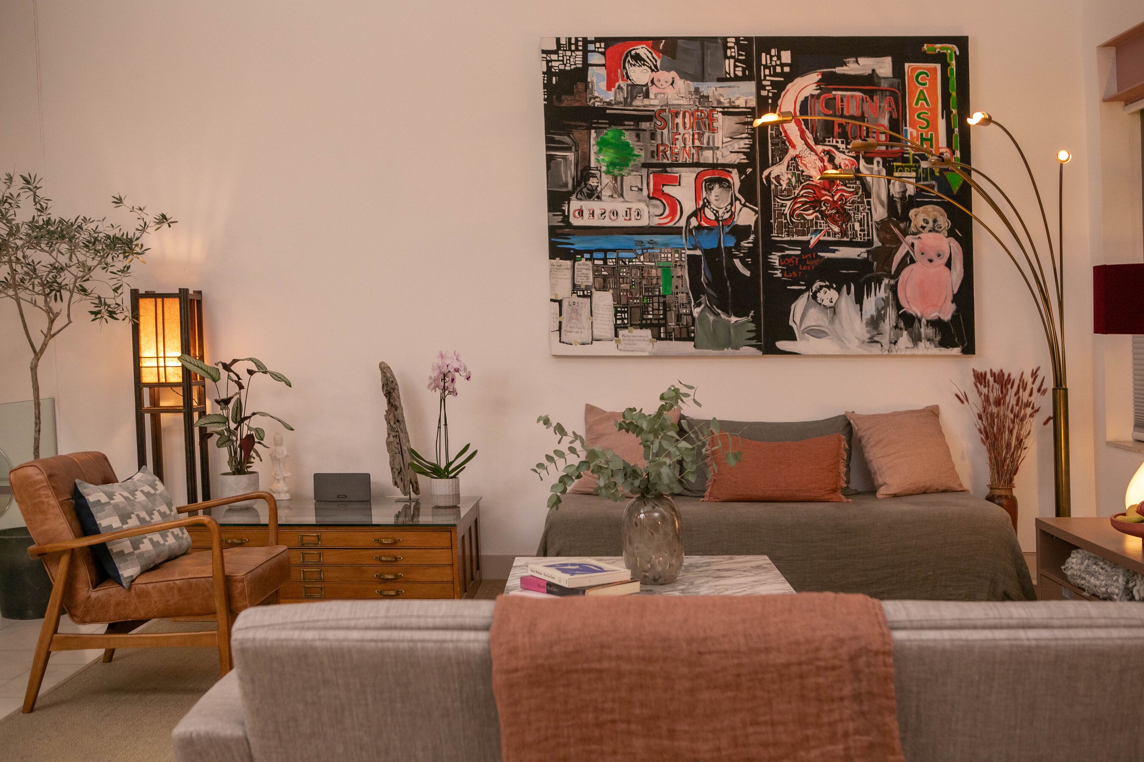 A modern bedroom with a large abstract painting, a bed with a grey and brown color scheme, a wooden side table, and a floor lamp.