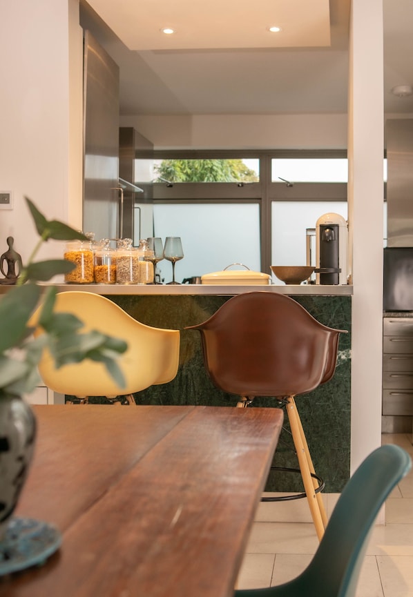 A modern kitchen with a dining area, featuring a marble countertop, a coffee machine, and a large window with a view of greenery. - The Luxury Inn (London)
