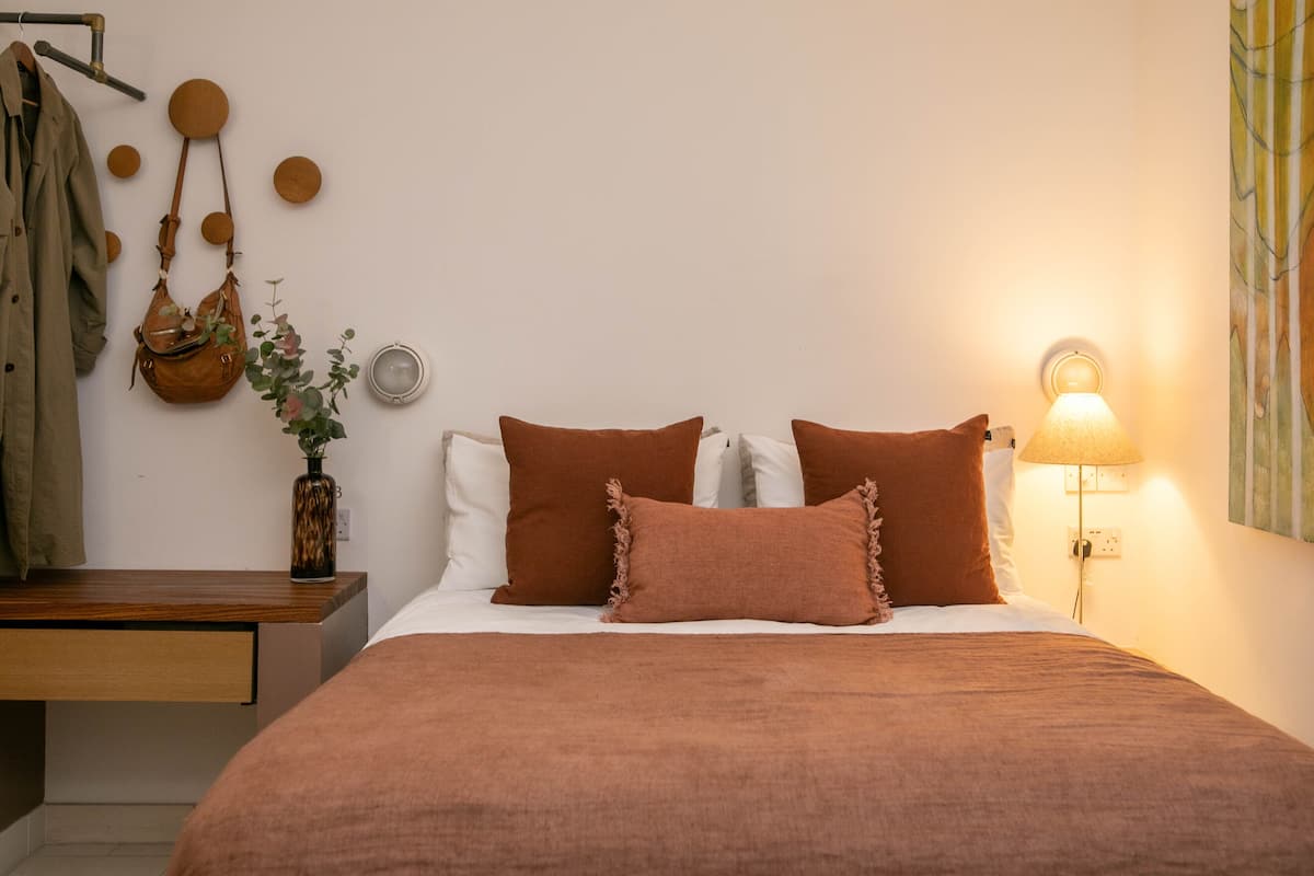 Un dormitorio con cama, almohadas, mesita de noche, lámpara, reloj en la pared y bolso colgado.