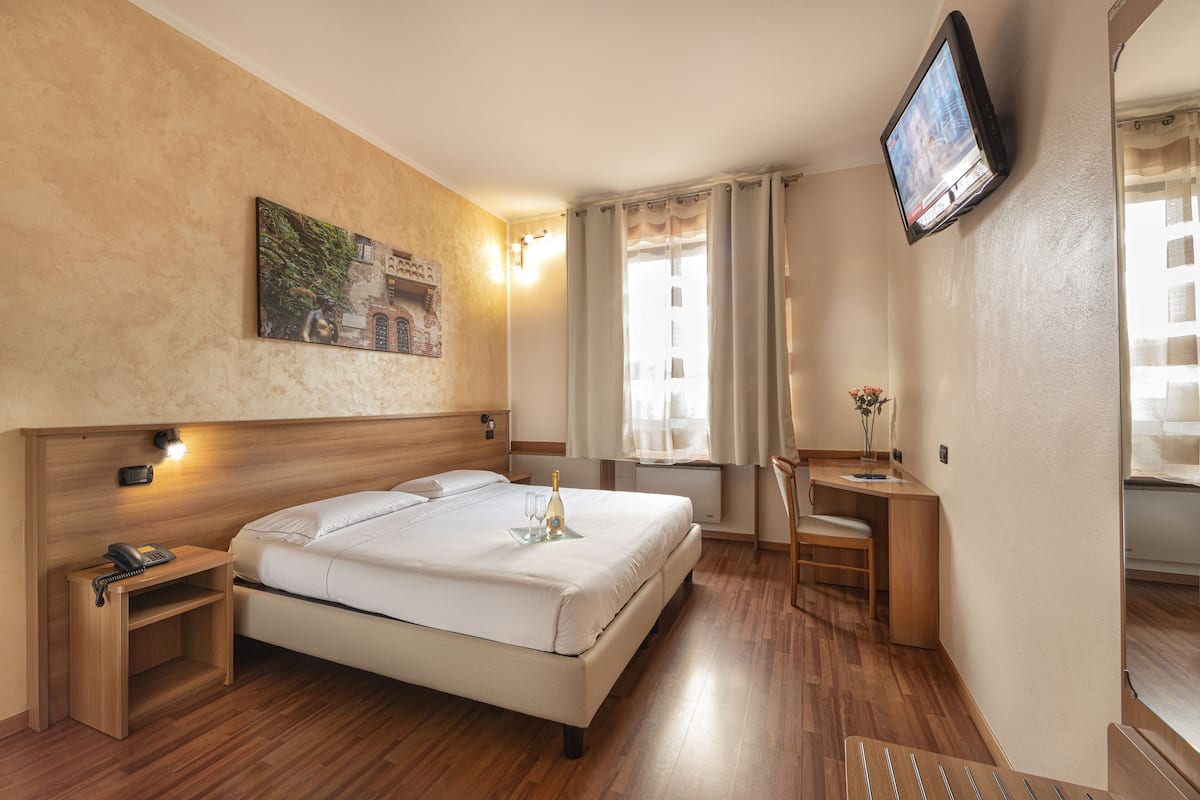 Une chambre d’hôtel comprenant un lit, une table de chevet avec un téléphone, un bureau avec une chaise, un téléviseur fixé au mur et une fenêtre avec des rideaux.