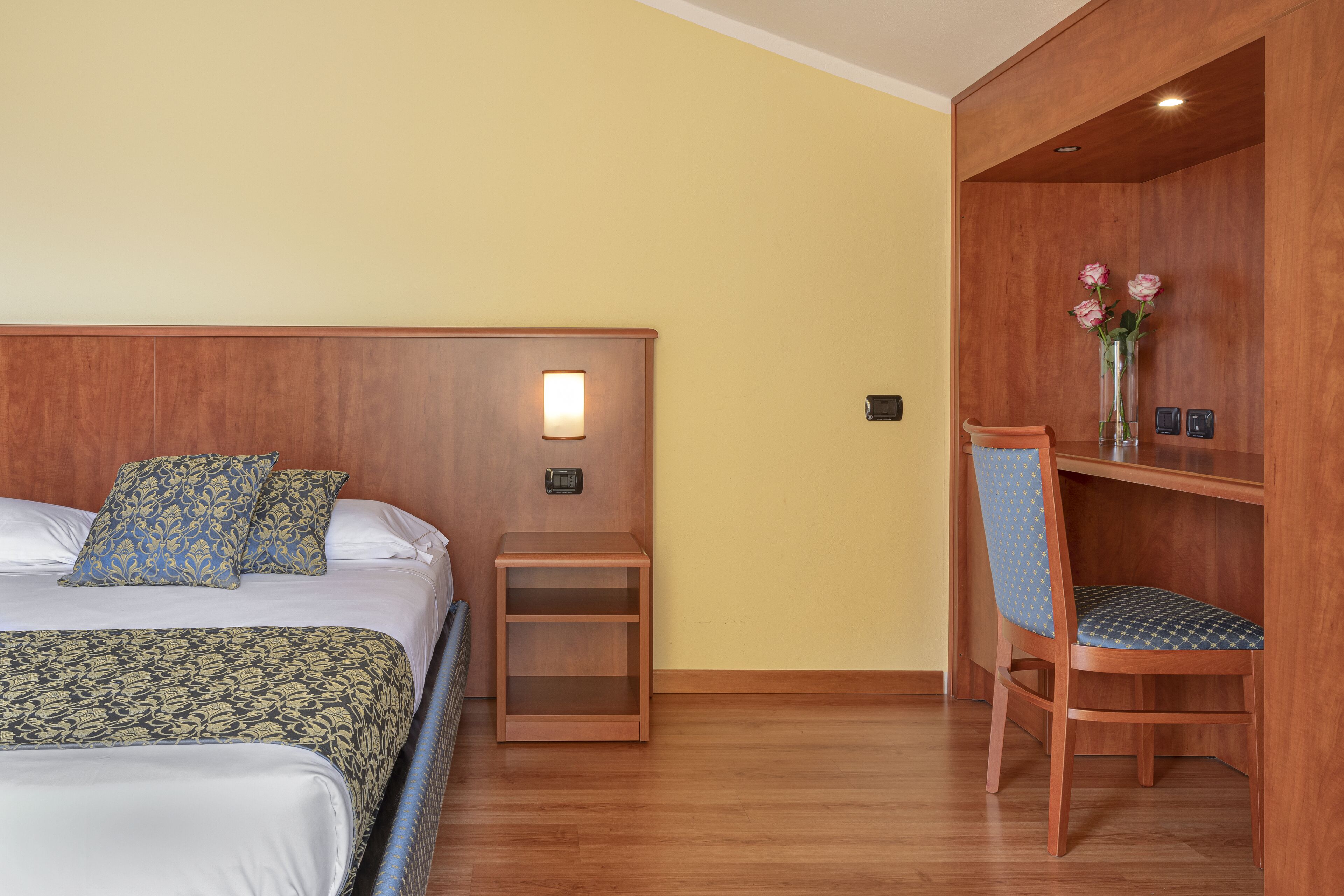 Een hotelkamer met twee bedden, een nachtkastje, een houten stoel en een klein plankje met bloemen.