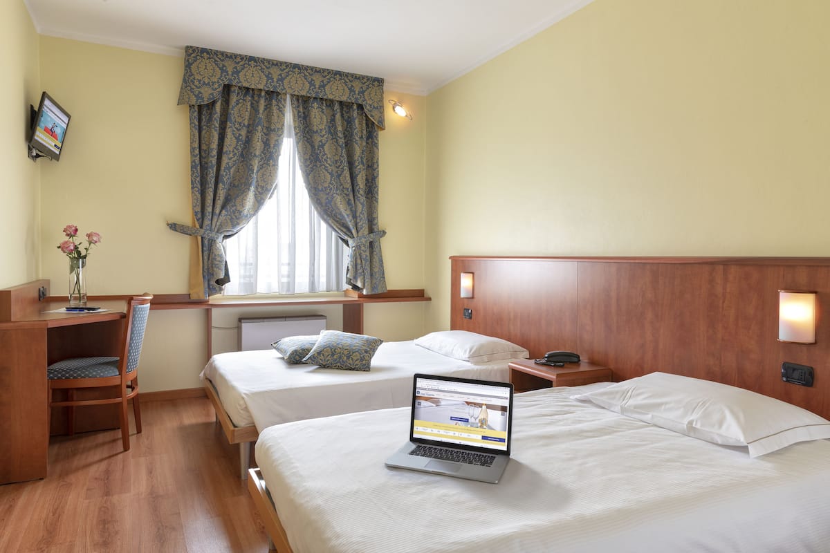 Une chambre d’hôtel avec deux lits, un bureau avec un ordinateur portable, une chaise et une fenêtre avec des rideaux.