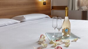 Ein ordentlich bezogenes Bett mit weißen Leinen, eine Champagnerflasche, zwei Gläser und verstreute Rosenblätter auf einem Glastablett.