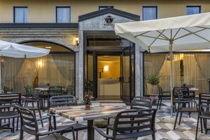 An outdoor dining area with tables and chairs, a potted plant, and a building entrance with a door and windows. - Hotel Postumia (Villafranca di Verona)