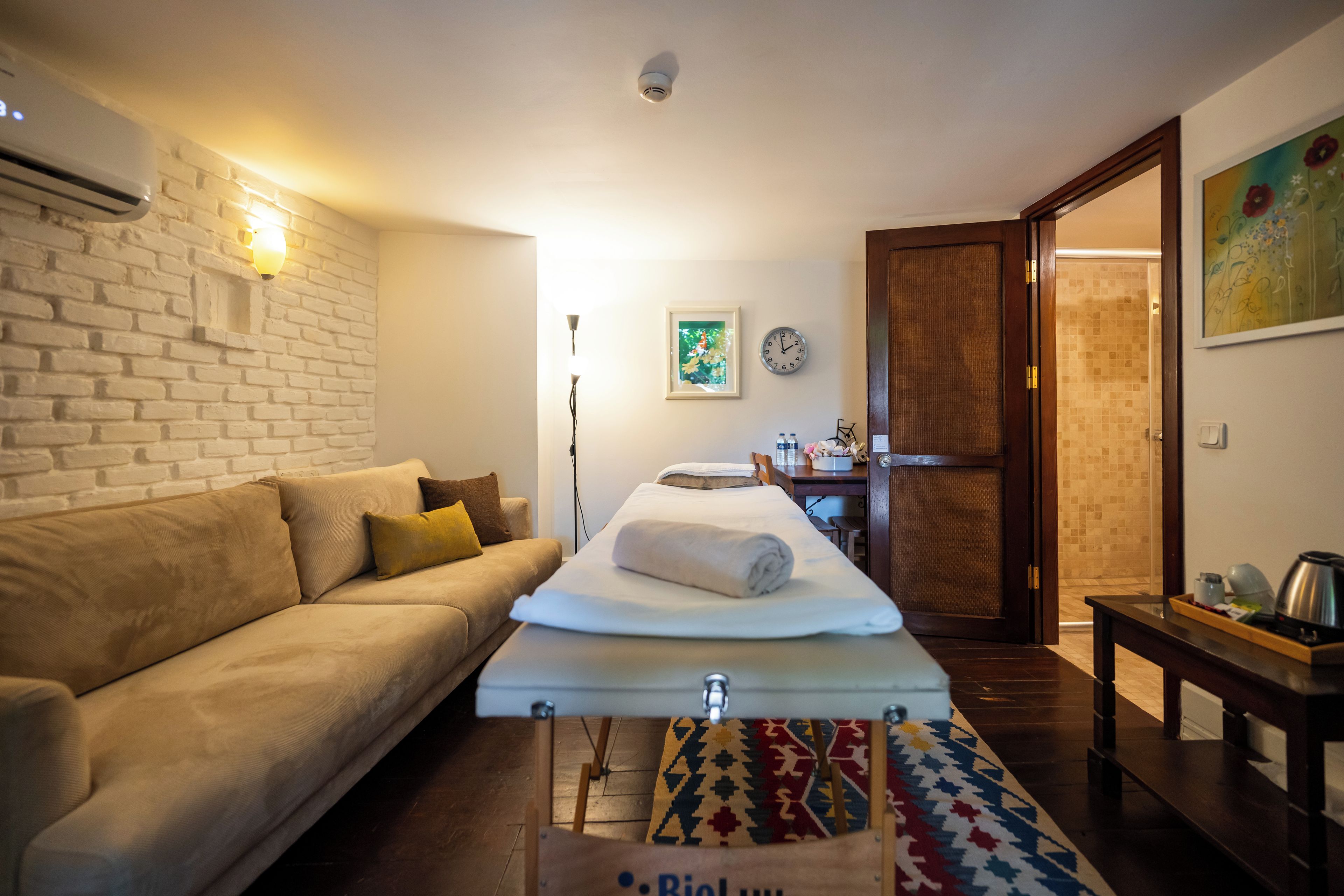 Quarto de hotel com sofá, cama com toalha enrolada, mesa de cabeceira, relógio, porta e ar-condicionado fixado na parede.