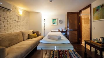 Quarto de hotel com sofá, cama com toalha enrolada, mesa de cabeceira, relógio, porta e ar-condicionado fixado na parede.